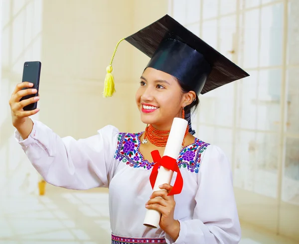 Young female student wearing traditional blouse with graduation hat, holding formal paper diploma roll while taking selfie using mobile phone, smiling to camera
