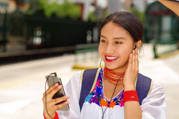 Pretty young woman wearing traditional andean blouse and blue backpack, waiting for bus at outdoors station platform, looking to mobile screen with headphones connected smiling happily