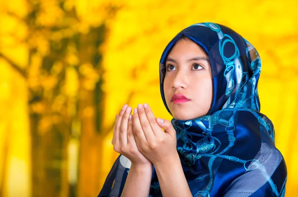 Beautiful young muslim woman wearing blue colored hijab, facing camera folding hands performing muslim prayer, autumn forest background