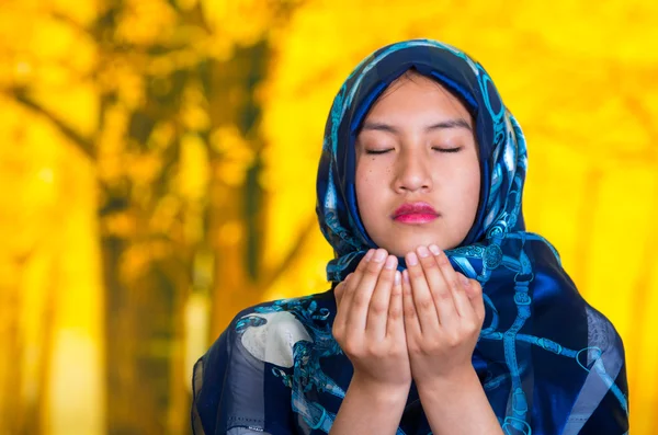 Beautiful young muslim woman wearing blue colored hijab, facing camera folding hands performing muslim prayer, autumn forest background