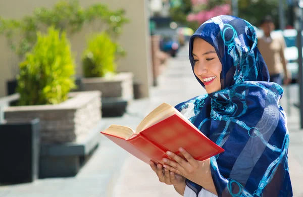 Beautiful young muslim woman wearing blue colored hijab, holding thick reed book and reading in street, outdoors urban background