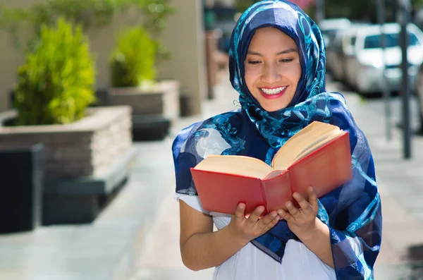 Beautiful young muslim woman wearing blue colored hijab, holding thick reed book and reading in street, outdoors urban background