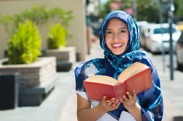 Beautiful young muslim woman wearing blue colored hijab, holding thick reed book and reading in street, outdoors urban background