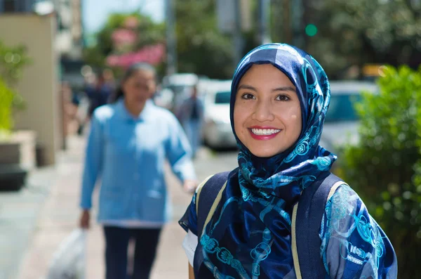 Beautiful young muslim woman wearing blue colored hijab and backpack, posing happily in street smiling to camera, outdoors urban background