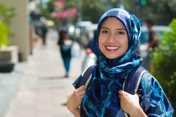 Beautiful young muslim woman wearing blue colored hijab and backpack, posing happily in street smiling to camera, outdoors urban background
