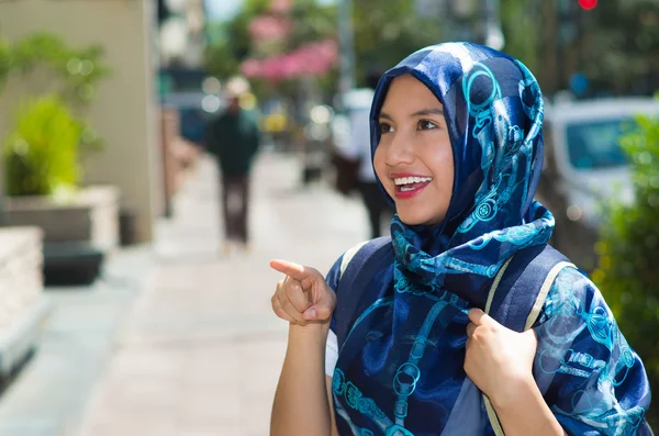Beautiful young muslim woman wearing blue colored hijab, pointing finger smiling, outdoors urban background