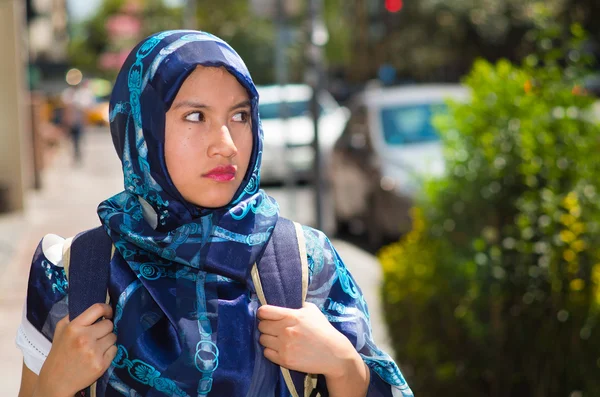 Beautiful young muslim woman wearing blue colored hijab and backpack, posing with thoughtful serious facial expression in street, outdoors urban background