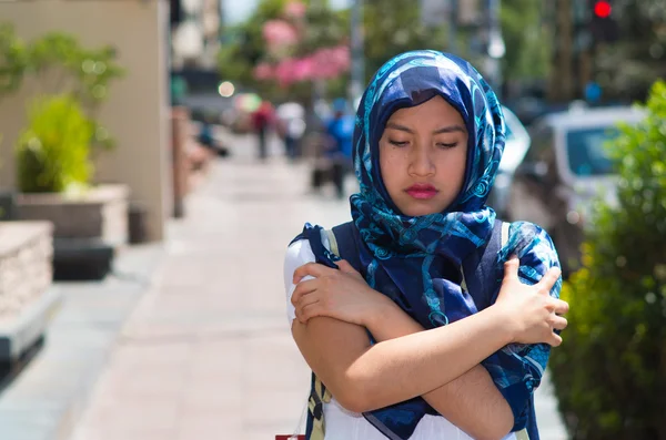 Beautiful young muslim woman wearing blue colored hijab and backpack,, interacting being cold rubbing arms using hands in street, serious facial expression, outdoors urban background
