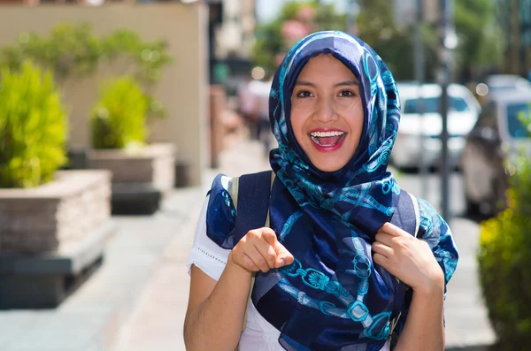 Beautiful young muslim woman wearing blue colored hijab, pointing finger smiling, outdoors urban background