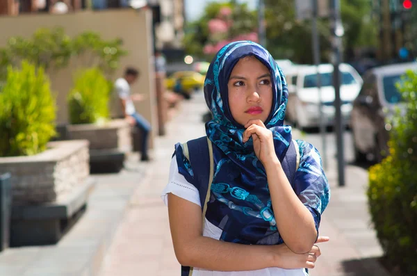 Beautiful young muslim woman wearing blue colored hijab and backpack, posing with thoughtful serious facial expression in street, outdoors urban background