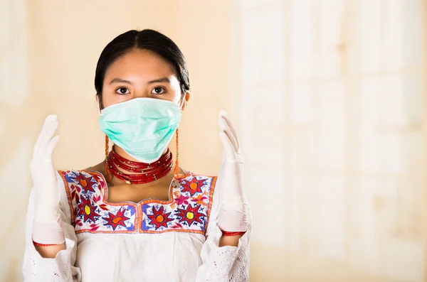 Young beautiful woman dressed in doctors coat and red necklace, face covered with facial mask looking into camera, egg white clinic background