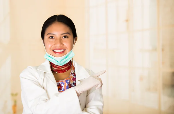 Young beautiful woman dressed in doctors coat and red necklace, facial mask pulled down to chin, smiling happily, egg white clinic background