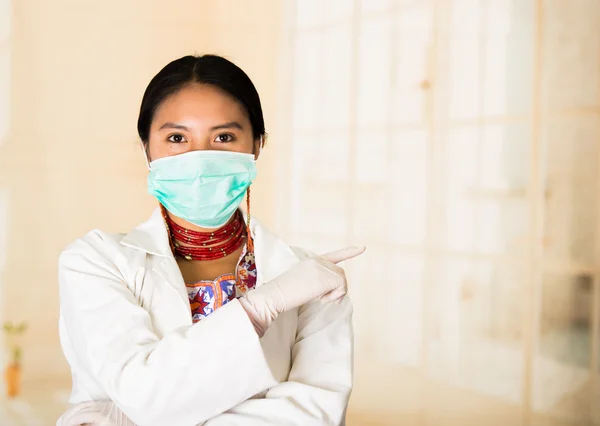 Young beautiful woman dressed in doctors coat and red necklace, face covered with facial mask looking into camera, egg white clinic background