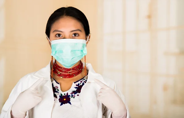 Young beautiful woman dressed in doctors coat and red necklace, face covered with facial mask looking into camera, egg white clinic background
