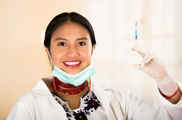 Young beautiful woman dressed in doctors coat and red necklace, facial mask pulled down to chin, holding up syringe smiling happily, egg white clinic background