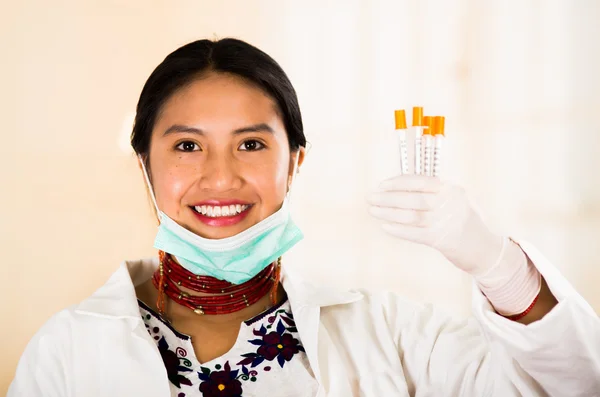 Young beautiful woman dressed in doctors coat and red necklace, facial mask pulled down to chin, holding up syringes smiling happily, egg white clinic background