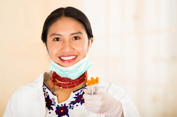 Young beautiful woman dressed in doctors coat and red necklace, facial mask pulled down to chin, holding up syringes smiling happily, egg white clinic background