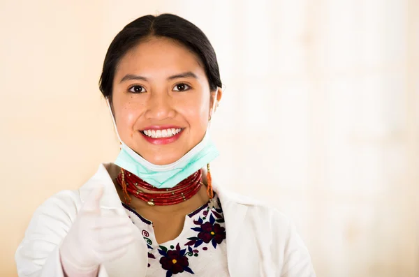 Young beautiful woman dressed in doctors coat and red necklace, facial mask pulled down to chin, smiling happily, egg white clinic background