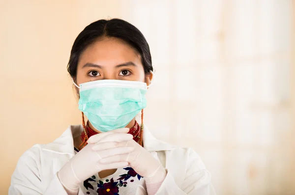 Young beautiful woman dressed in doctors coat and red necklace, face covered with facial mask looking into camera, egg white clinic background