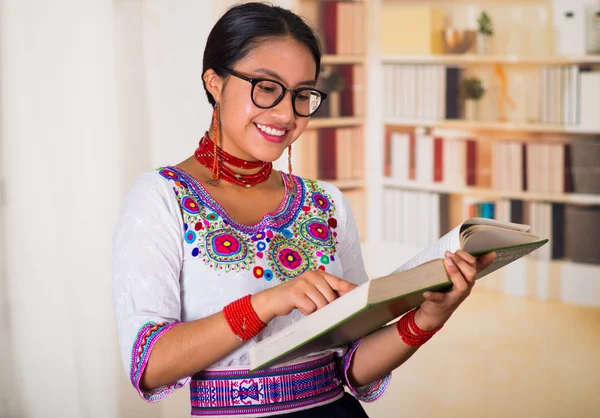 Beautiful young lawyer wearing traditional andean blouse and glasses, holding book reading, bookshelves background