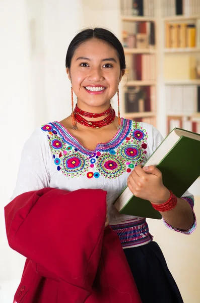 Beautiful young lawyer wearing traditional andean blouse, holding red jacket and green book smiling to camera, bookshelves background