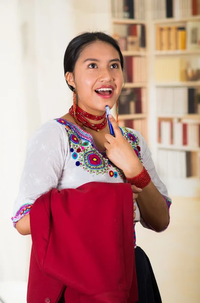 Beautiful young lawyer wearing traditional andean blouse, holding red jacket smiling to camera, bookshelves background