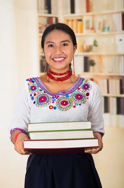 Beautiful young lawyer wearing traditional andean blouse and red necklace, holding stack of books, smiling to camera, bookshelves background