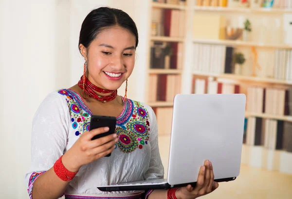 Beautiful young lawyer wearing traditional andean blouse and red necklace, holding laptop looking at mobile screen smiling, bookshelves background