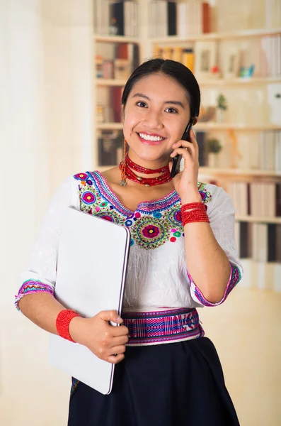 Beautiful young lawyer wearing traditional andean blouse and red necklace, holding laptop talking on phone smiling, bookshelves background