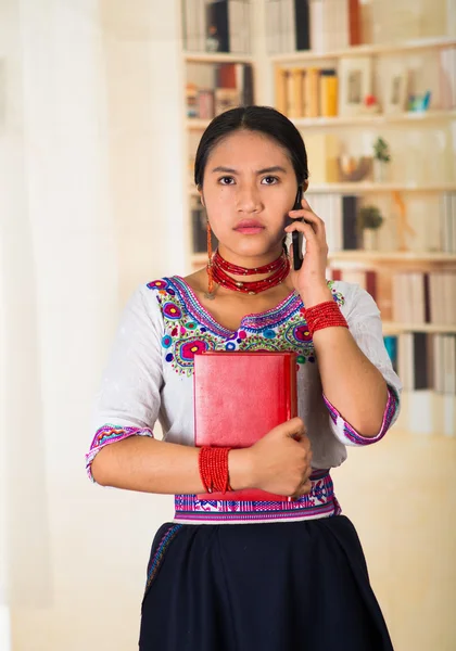 Beautiful young lawyer wearing black skirt, traditional andean blouse with necklace, standing posing for camera, holding red book and talking on phone, serious facial expression, bookshelves