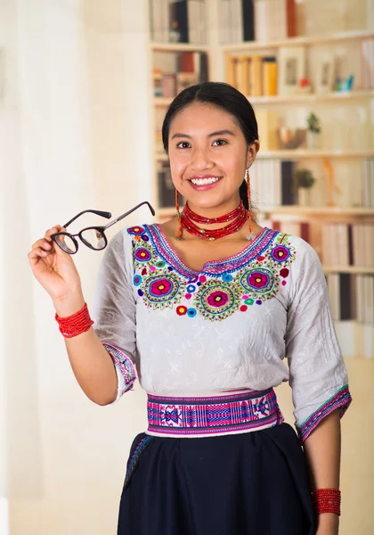 Beautiful young lawyer wearing black skirt, traditional andean blouse with necklace, standing posing for camera, holdig glasses, smiling happily, bookshelves background