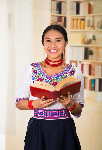 Beautiful young lawyer wearing black skirt, traditional andean blouse with necklace, standing posing for camera, holding red book reading happily, bookshelves background.