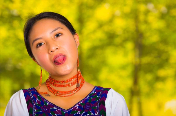 Headshot beautiful young woman wearing traditional andean blouse with red necklace, posing for camera holding tongue out, green forest background