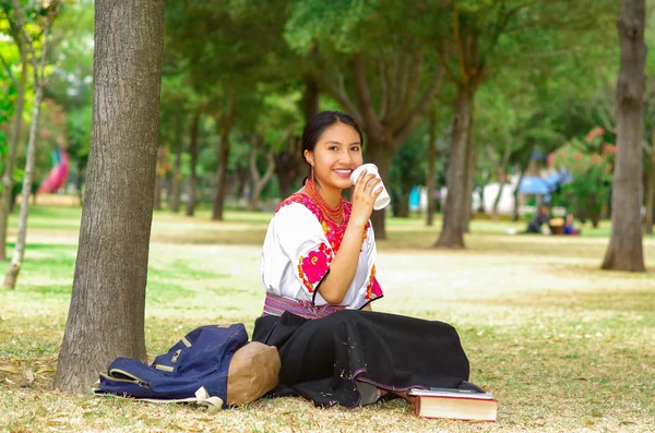 Young woman wearing traditional andean skirt and blouse with matching red necklace, sitting on grass next to tree in park area, relaxing while drinking coffee smiling happily