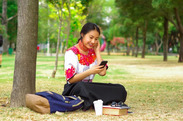 Young woman wearing traditional andean skirt and blouse with matching red necklace, sitting on grass next to tree in park area, relaxing while using mobile phone headphones connected, smiling happily