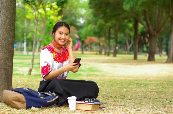 Young woman wearing traditional andean skirt and blouse with matching red necklace, sitting on grass next to tree in park area, relaxing while using mobile phone headphones connected, smiling happily