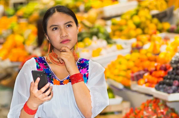 Beautiful young hispanic woman wearing andean traditional blouse using mobile phone inside fruit market, thoughtful facial expression, colorful healthy food selection in background