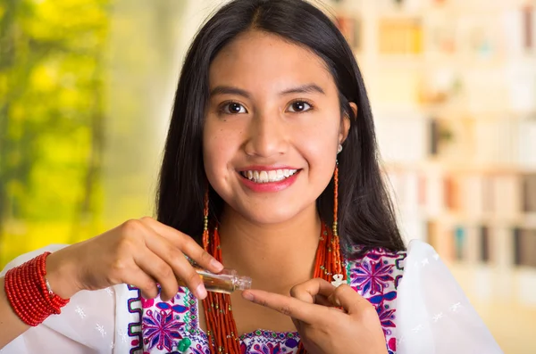 Beautiful hispanic woman wearing white blouse with colorful embroidery, applying cosmetic product to finger during makeup routine, smiling happily, garden background