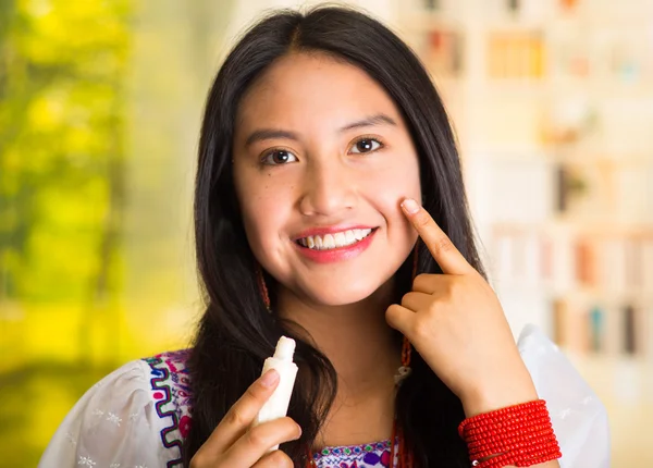 Beautiful hispanic woman wearing white blouse with colorful embroidery, applying cream onto face using finger during makeup routine, smiling happily, garden background