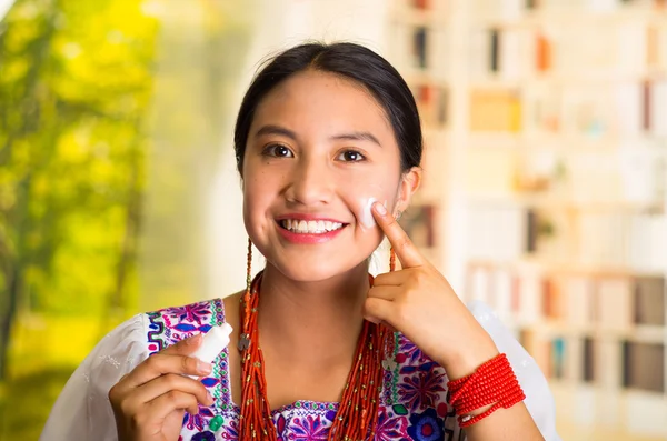 Beautiful hispanic woman wearing white blouse with colorful embroidery, applying cream onto face using finger during makeup routine, smiling happily, garden background