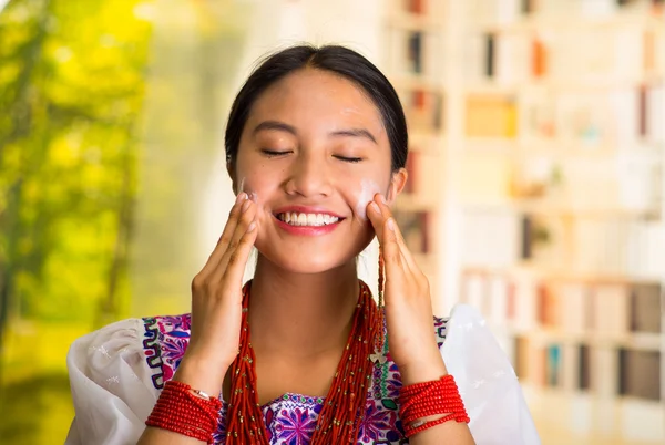 Beautiful hispanic woman wearing white blouse with colorful embroidery, applying cream onto face using both hands during makeup routine, smiling happily, garden background
