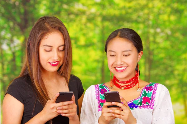 Two beautiful young women posing for camera, one wearing traditional andean clothing, the other in casual clothes, both pressing phones looking at mobile screens, park background