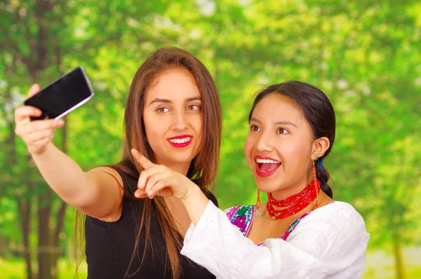 Two beautiful young women standing together facing camera, one wearing traditional andean clothing, the other in casual clothes, holding up mobile posing for selfie smiling, park background
