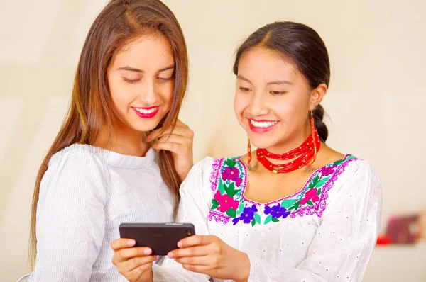 Two beautiful young women posing for camera, one wearing traditional andean clothing, the other in casual clothes, holding mobile between them interacting, both smiling, park background