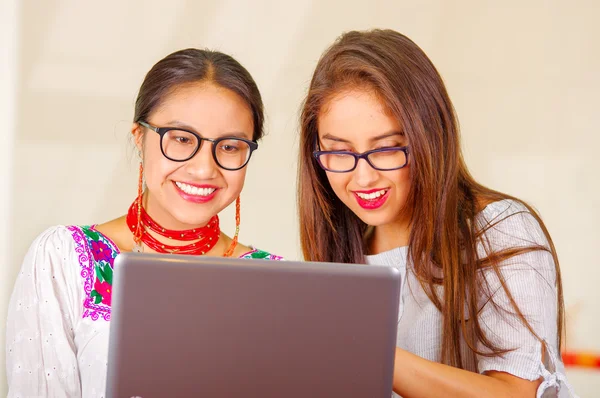 Two beautiful young women posing for camera, one wearing traditional andean clothing, the other in casual clothes, holding laptop between them interacting looking at screen, both smiling, park
