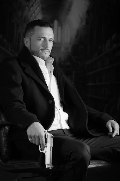 Elegant man sitting in a chair holding  gun over dark background