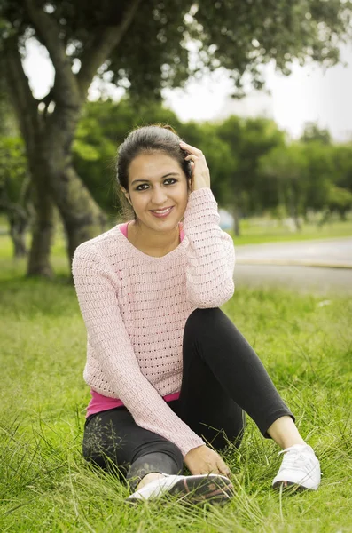 Hispanic brunette sitting on grass in yoga clothing left knee bent and touching hair with arm while looking into camera smiling