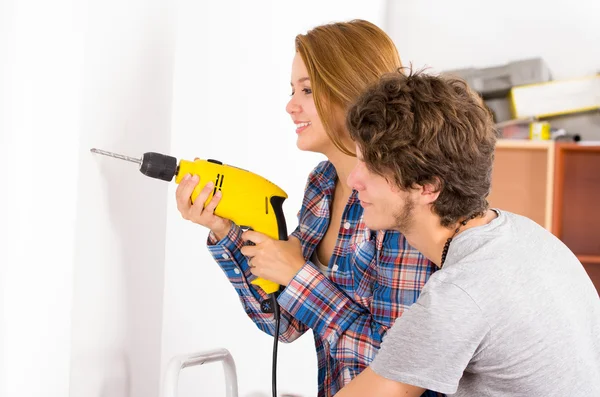 Couple renovating together as woman using power drill on wall with man standing next to her observing