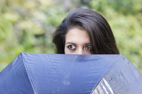 Closeup good-looking hispanic brunette woman head peeking up from behind black umbrella revealing only her eyes with outdoors park background blurry