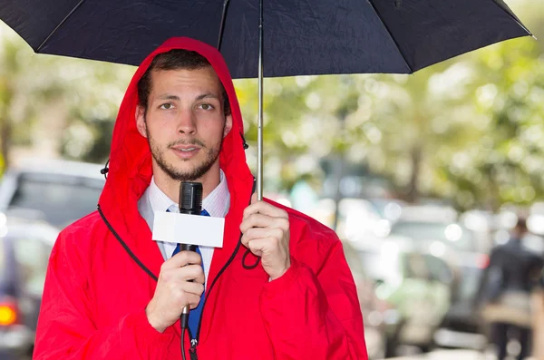 Successful handsome male journalist wearing red rain jacket working in rainy weather outdoors park environment holding microphone and umbrella, live broadcasting
